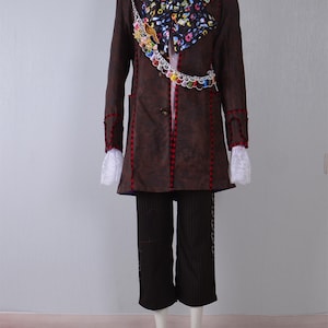 May include: A brown coat with red and white trim, a white shirt with a pink bow tie, a black and white patterned scarf, and a chain with colorful charms. The coat has a pocket on the left side. The pants are black with white stripes.