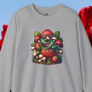 Peut inclure: Un sweat-shirt gris avec une grenouille verte assise sur une fraise rouge et entourée de champignons. La grenouille porte un chapeau de champignon rouge.