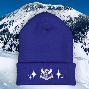 May include: A royal blue beanie with a white embroidered design featuring a dragon, an open book, and starbursts. The beanie is made of a soft, knitted material, perfect for cold weather. The background shows a snowy mountain.