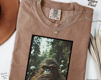 May include: Espresso-colored t-shirt with a graphic of a Bigfoot-like creature and aliens in a forest setting. The shirt has a crew neck and a Comfort Colors label. A silver necklace with a small pendant rests on the shirt.