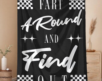 May include: Black blanket with white text that reads "FART AROUND AND FIND OUT." The blanket has a checkered pattern at the top and bottom, and star shapes between the words. The blanket is held up by a person.