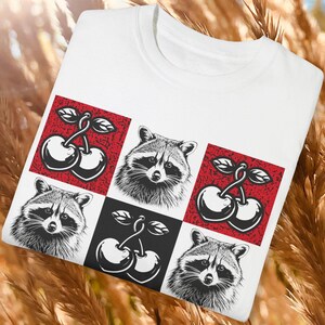 May include: White t-shirt featuring a grid design with raccoons and cherries. The design includes black and white raccoons and cherries on red and black squares. The shirt has a crew neck and short sleeves.