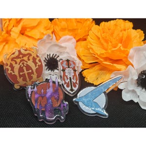 May include: Five colorful enamel pins with glitter accents. The pins feature different designs, including a castle, a shield with a cross, a shield with a bat, a shield with a flower, and a blue and white abstract design.