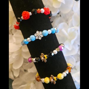 Puede incluir: Cuatro pulseras de cuentas con diferentes dijes. La primera pulsera tiene flores rojas y cuentas negras. La segunda pulsera tiene cuentas azules y un dije de copo de nieve. La tercera pulsera tiene cuentas negras, blancas y rosadas con un dije de abeja. La cuarta pulsera tiene cuentas amarillas y un dije de abeja.