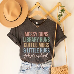 May include: A dark grey t-shirt with the text "Messy Buns Library Runs Coffee Mugs & Little Hugs #homeschool" in colourful text. The shirt is hanging on a wooden hanger with a straw hat and a woven bag.