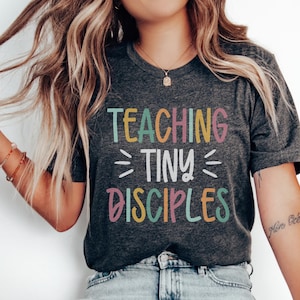 May include: Dark gray t-shirt with the text "TEACHING TINY DISCIPLES" in colorful letters. The shirt is paired with light wash denim jeans and white slip-on shoes. The overall style is casual and comfortable.