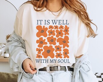 May include: A white t-shirt with the text "It is well with my soul" and a graphic of orange flowers with blue centers.