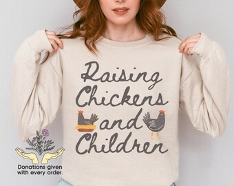 May include: A beige long-sleeved sweatshirt with the text "Raising Chickens and Children" in black script. The text is accompanied by a black and white illustration of a chicken in a nest and a chicken standing on two legs. The text "Donations given with every order." is printed below the illustration.