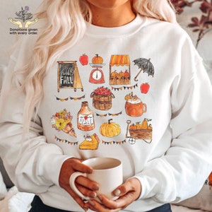 May include: White sweatshirt with a fall-themed design featuring pumpkins, apples, a pie, a coffee mug, and a sign that says "hello fall".