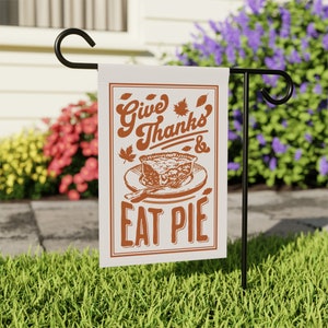 May include: A rectangular garden flag with the text "Give Thanks & Eat Pie" in brown lettering. The flag features a pie graphic and maple leaf accents. The flag is displayed on a black metal stand in a grassy yard.