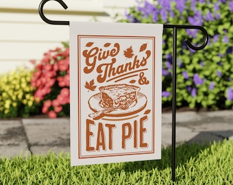 May include: A rectangular garden flag with the text "Give Thanks & Eat Pie" in brown lettering. The flag features a pie graphic and maple leaf accents. The flag is displayed on a black metal stand in a grassy yard.