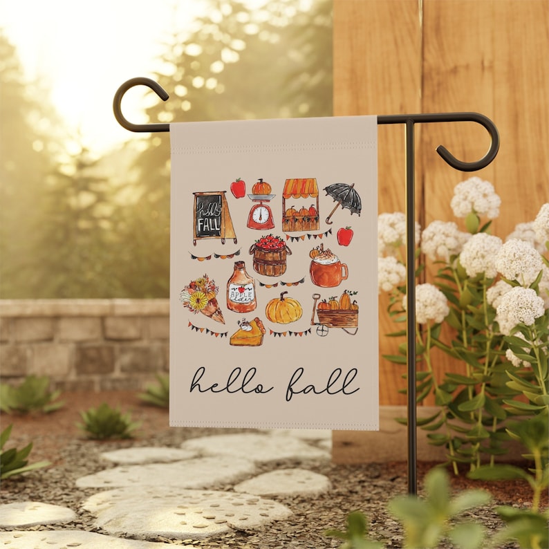May include: A beige garden flag featuring a watercolour illustration of autumn elements, such as pumpkins, apples, and a chalkboard sign that reads "Hello Fall." The flag is displayed on a black metal stand in a garden.