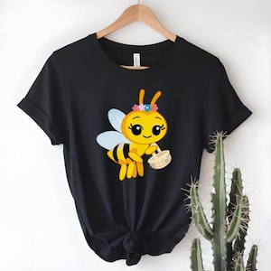 Peut inclure: T-shirt noir avec une abeille de dessin anim&eacute; portant une couronne de fleurs et un panier. L'abeille est jaune et noire avec des ailes bleues. Le t-shirt est suspendu &agrave; un cintre en bois.