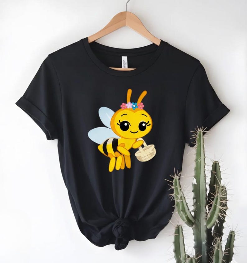 Peut inclure: T-shirt noir avec une abeille de dessin anim&eacute; portant une couronne de fleurs et un panier. L'abeille est jaune et noire avec des ailes bleues. Le t-shirt est suspendu &agrave; un cintre en bois.