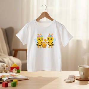 Peut inclure: T-shirt blanc avec une illustration de dessin anim&eacute; de deux abeilles jaunes avec des couronnes de fleurs et des ailes bleues, tenant un panier. Les abeilles sont centr&eacute;es sur le t-shirt. Le t-shirt est suspendu &agrave; un cintre en bois.