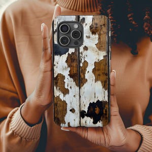 May include: A smartphone case with a distressed wood design. The case features a pattern of brown wood with white and black paint splatters. The case has a camera module with three lenses.