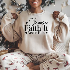 May include: A beige sweatshirt with the text "Choose Faith It Never Fails" in black script. The sweatshirt is paired with patterned pajama pants. The image is well-lit, showcasing the details of the clothing.