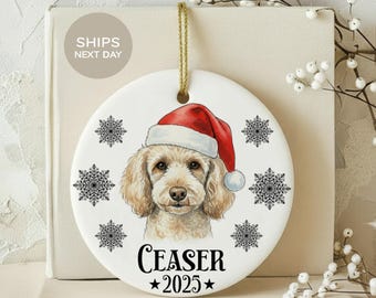 May include: A white ceramic Christmas ornament featuring a dog wearing a Santa hat. The ornament has the name "Ceaser" and the year "2025" printed below the dog's image. Black snowflake designs are around the dog's image.