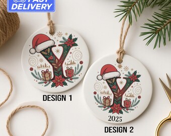 May include: Two white ceramic Christmas ornaments with the letter "Y" in a festive design. Each ornament features a Santa hat, poinsettias, gifts, and the text "DESIGN 1" and "2025 DESIGN 2". They hang from twine.