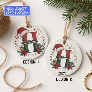 May include: Two white ceramic Christmas ornaments with the letter "H" in a festive design. One ornament says "Design 1" and the other says "2025 Design 2". Each ornament has a red Santa hat, green and red floral accents, and a twine loop for hanging.