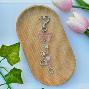 May include: A pink and silver charm keychain with a bow, hearts, and a cat charm. The keychain is hanging from a wooden tray.