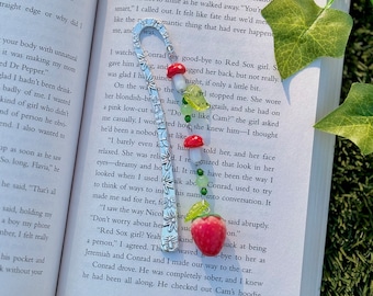 May include: A silver bookmark with a strawberry charm and green and red glass beads. The bookmark is open to a page in a book with the text "The Summer I Turned Pretty" and the page number 121.