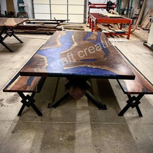 May include: A large wooden dining table with a blue river-like design in the center. The table has a dark wood finish and is supported by a black metal base. Matching benches with black metal legs are also present.
