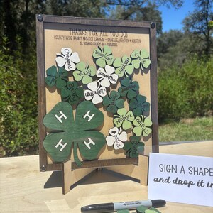 May include: A wooden display board featuring a large green four-leaf clover with the 4-H emblem, surrounded by smaller clover-shaped cutouts with names. Text reads: THANKS FOR ALL YOU DO! COUNTY WIDE RABBIT PROJECT LEADERS - DANELLE, ASHTON & KIRSTIN.