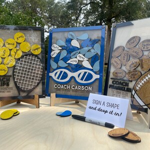 May include: Three wooden signs with a clear acrylic overlay, each featuring a different sport theme. The signs have a light wood background and are decorated with a tennis racket, swimming goggles, and a football. Each sign has a title with the name of a coach and the team name. The signs are on small wooden stands and there is a sign in the front that says "Sign a shape and drop it in!"