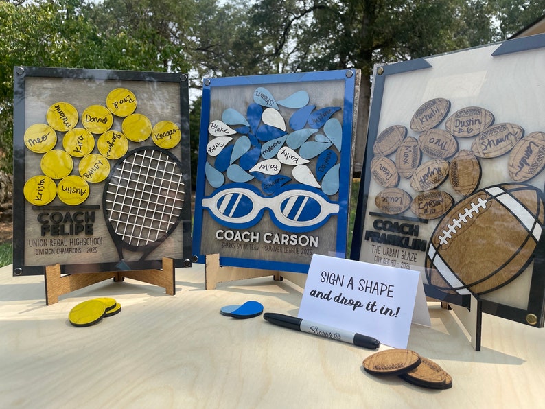 May include: Three wooden signs with a clear acrylic overlay, each featuring a different sport theme. The signs have a light wood background and are decorated with a tennis racket, swimming goggles, and a football. Each sign has a title with the name of a coach and the team name. The signs are on small wooden stands and there is a sign in the front that says "Sign a shape and drop it in!"