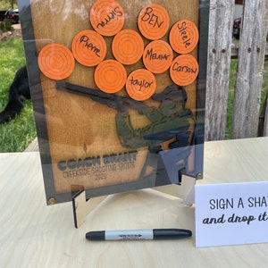 May include: A wooden sign featuring a silhouette of a person shooting a shotgun. The sign has orange targets with names written on them, including "Pierre", "BEN", "Steele", "Melame", "Carson", and "Taylor". The sign also reads "COACH BRANT", "CREEKSIDE SHOOTING SPORTS", and "2025".