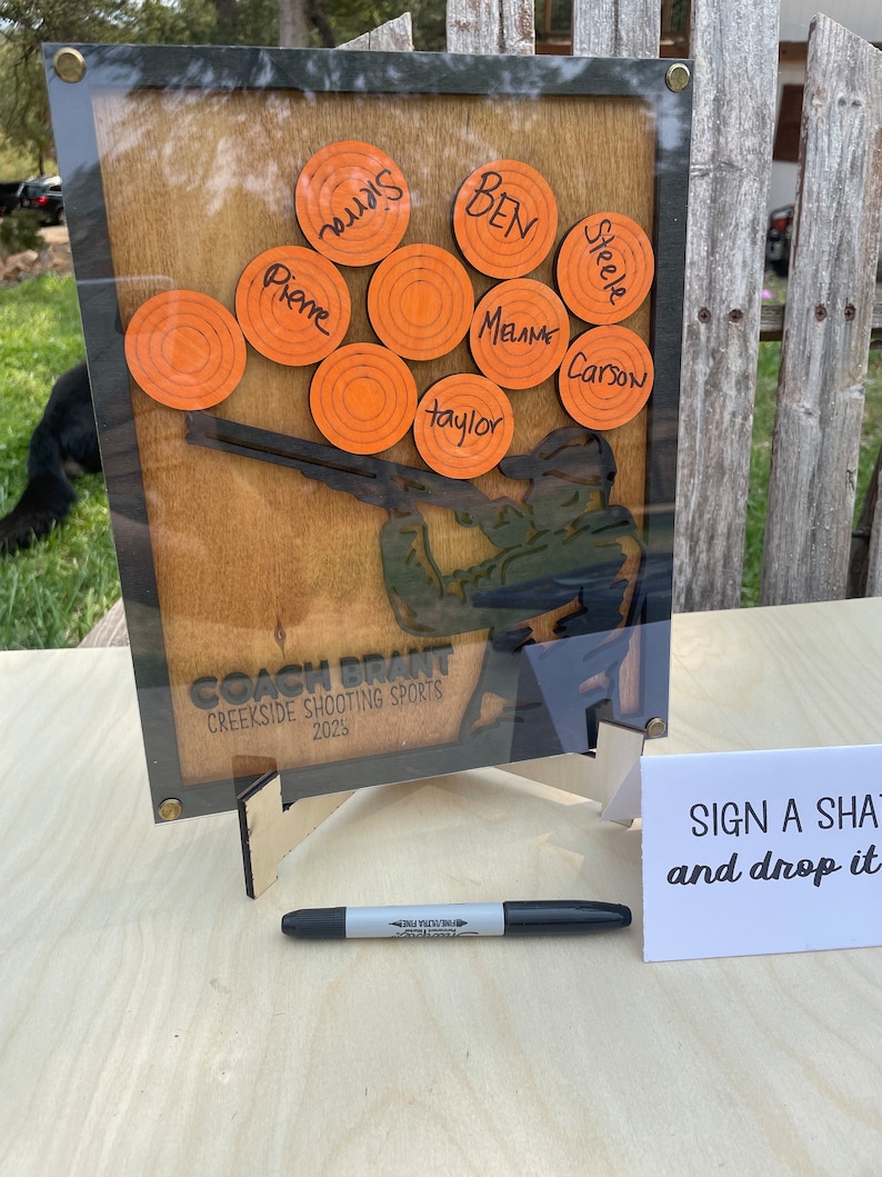 May include: A wooden sign featuring a silhouette of a person shooting a shotgun. The sign has orange targets with names written on them, including "Pierre", "BEN", "Steele", "Melame", "Carson", and "Taylor". The sign also reads "COACH BRANT", "CREEKSIDE SHOOTING SPORTS", and "2025".
