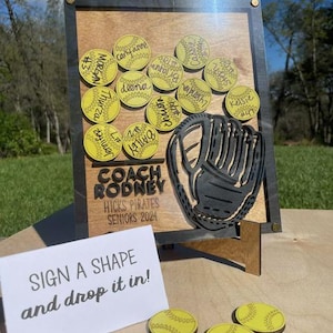 May include: A wooden sign with a black and gray softball glove cutout. The sign says "Coach Rodney Hicks Pirates Seniors 2024". There are yellow softball-shaped pieces with names written on them. A sign below says "Sign a shape and drop it in!"