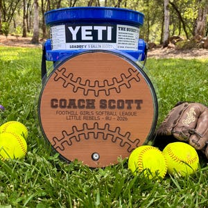 May include: A blue Yeti LoadOut 5-gallon bucket with a wooden softball-themed plaque that reads "COACH SCOTT" and "FOOTHILL GIRLS SOFTBALL LEAGUE LITTLE REBELS - 8U - 2026". The image includes yellow softballs and a baseball glove.