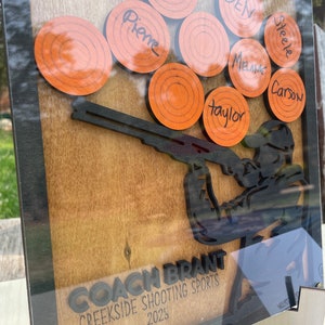 May include: A wooden plaque featuring a silhouette of a person holding a shotgun. The plaque has a brown background and black lettering. The lettering reads "Coach Brant Creekside Shooting Sports 2023". There are nine orange clay targets with names written on them.