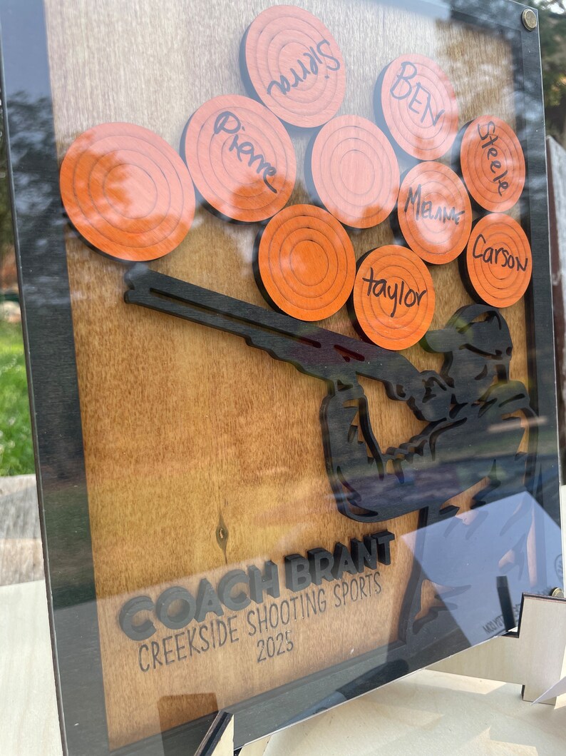 May include: A wooden plaque featuring a silhouette of a person holding a shotgun. The plaque has a brown background and black lettering. The lettering reads "Coach Brant Creekside Shooting Sports 2023". There are nine orange clay targets with names written on them.