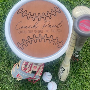 May include: A wooden round sign with a baseball design and the text "Coach Paul" and "Foothill Girls Softball - 10U - Fall 2024". The sign is on a white background and surrounded by green grass. There are two baseball bats and a baseball glove in the image.