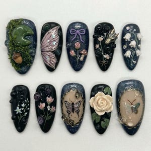 Op de afbeelding: Een verzameling van tien kunstnagels met diverse ontwerpen. De ontwerpen omvatten een waterlelie, elfen vleugels, een strik, een wolf, bloemen, een vlinder en een roos. De nagels zijn geschilderd in donkerblauw, zwart en beige met glitteraccenten.