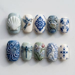 May include: A set of ten oval-shaped press-on nails featuring various raised and painted designs including a white seashell, a blue bird, blue and white floral patterns, a crab, an octopus, a dragonfly, flowers, and a white rabbit.