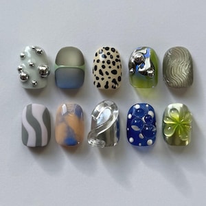 Può includere: Dieci unghie finte con vari design. Le unghie presentano colori come argento, blu, verde e beige, con motivi a pois, vortici e abbellimenti 3D. Alcune unghie hanno accenti metallici, offrendo una varietà di stili.