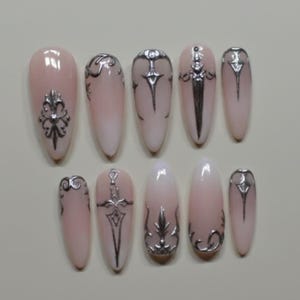 May include: A set of ten almond-shaped false nails. Each nail is painted with a light pink gradient and adorned with silver-colored embellishments. The designs include ornate patterns and sword motifs, creating a gothic or fantasy aesthetic.