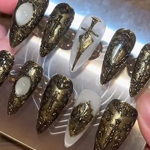 May include: A set of ornate, long, pointed artificial nails. The nails feature a metallic gold and bronze color scheme with intricate embossed designs. Some nails have a clear base with gold embellishments, including a sword and shield.