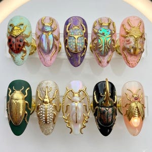 May include: A collection of ten artificial nails, each adorned with a unique, ornate beetle design. The nails feature a variety of colors, including green, pink, purple, and gold, with intricate details and textures. The beetles are the focal point, adding a touch of nature-inspired elegance.