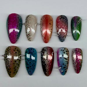 May include: Ten artificial nails in various colors, including pink, green, and red, with a speckled design. Each nail has a silver spike accent. The nails are arranged on a white surface.