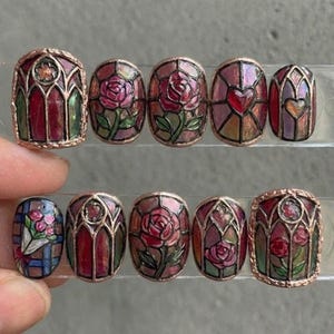 May include: A collection of ten press-on nails with a stained glass effect. The nails showcase rose and heart motifs, along with geometric patterns, in hues of red, pink, and green. Each nail is outlined with a copper-toned border.