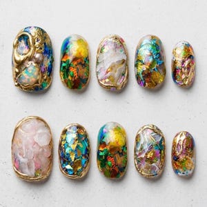 May include: A set of ten artificial nails with a gold trim. The nails feature a variety of designs, including iridescent flakes, opalescent accents, and gold leaf. The color palette includes blues, greens, yellows, and pinks.