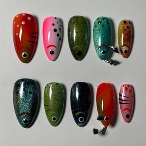 May include: A set of ten artificial nails, each painted with a unique fish-inspired design. Colors include red, pink, green, and blue, with details like dots, stripes, and metallic accents. Some nails feature small, silver-colored embellishments.