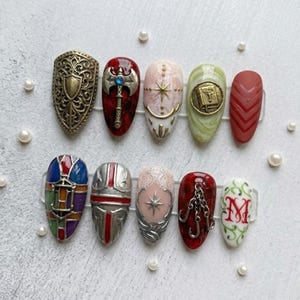 May include: A set of ten artificial nails with diverse designs. The designs include a shield, an axe, a star, a book, and a chevron pattern. Other nails feature a stained glass design, a helmet, a star, and a chain. The last nail has the letter "M".
