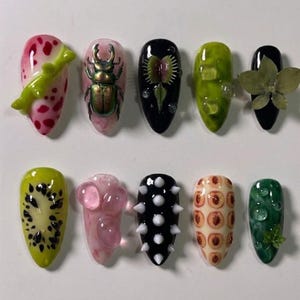 May include: A collection of ten uniquely designed artificial nails. The nails feature various designs, including a beetle, a Venus flytrap, and fruit slices. Colors include pink, green, black, and white. Some nails have 3D elements, such as a branch and spikes.