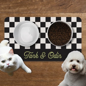 May include: A black and white checkered pet feeding mat with two white ceramic bowls. One bowl is filled with dry dog food and the other is empty. The mat has the text "Tank & Odin" in green lettering.